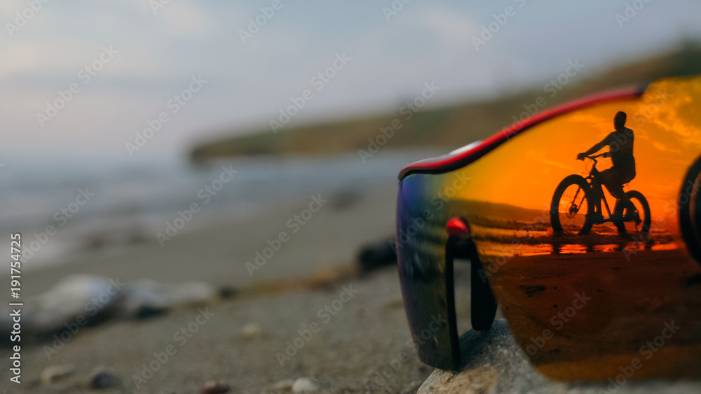 Naklejka premium Fat bike also called fatbike or fat-tire bike in summer driving on the beach. Sportsman is passing by in the reflection of sports glasses silhouetted against a beautiful sunset at the sea.