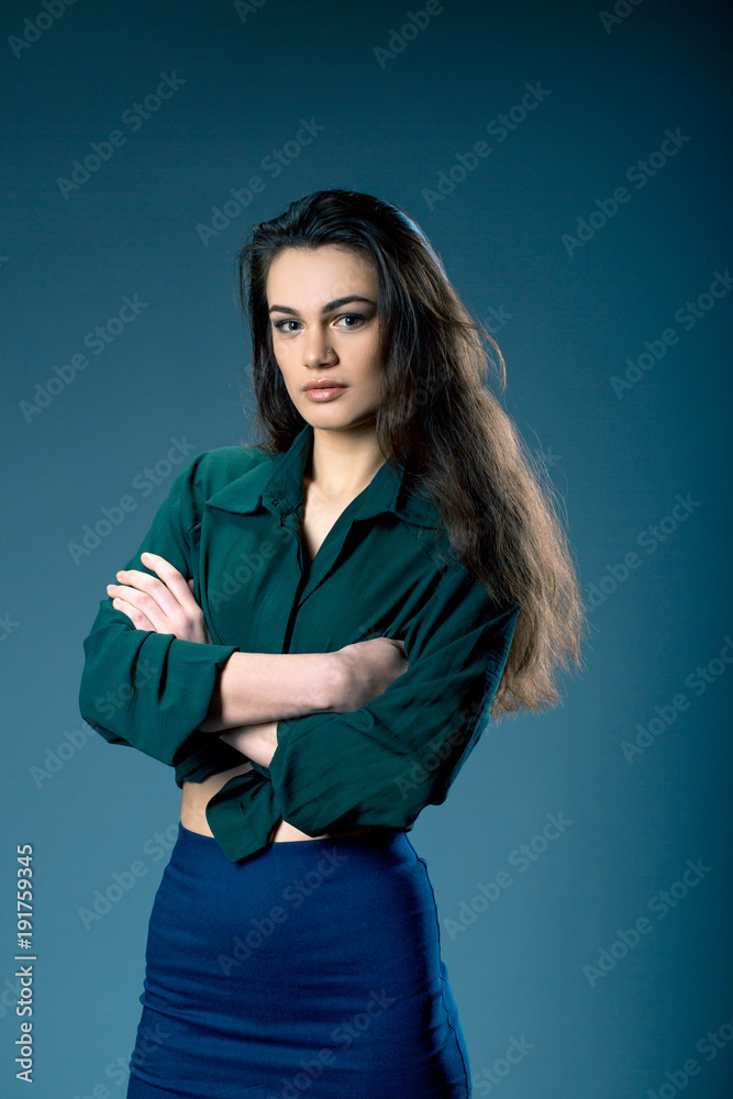 Portrait of young gorgeous dark-haired girl with provocative make up wearing high waisted blue skirt and green a shirt tied on the belly on blue grey background. Studio shot Portrait of young gorgeous dark-haired girl with provocative make up wearing high waisted blue skirt and green a shirt tied on the belly on blue grey background. Studio shot