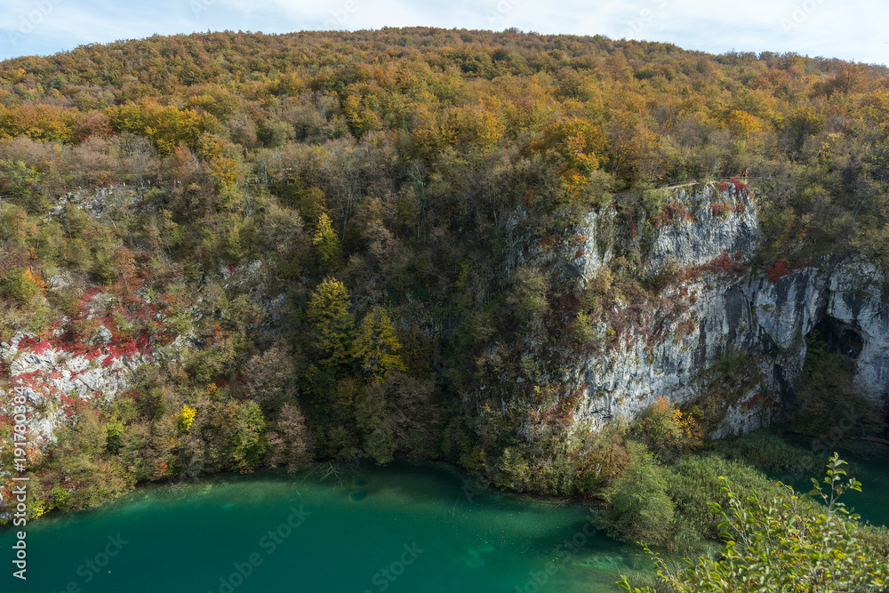 A gigantic white cliff covered in colorful trees in Plitvice Lakes National Park. The water is a turquoise green with clouds above.