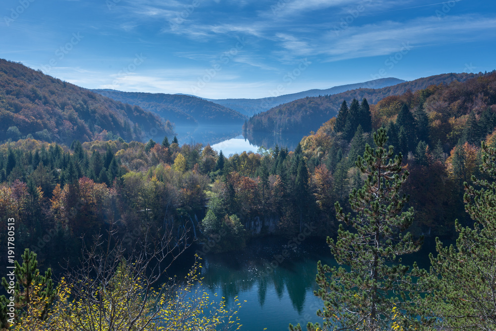 Two lakes are shown in Plitvice Lakes National Park. The trees are changing colors from green to yellow, orange and red. There is some fog above the lake and few clouds in the blue sky. Great distance