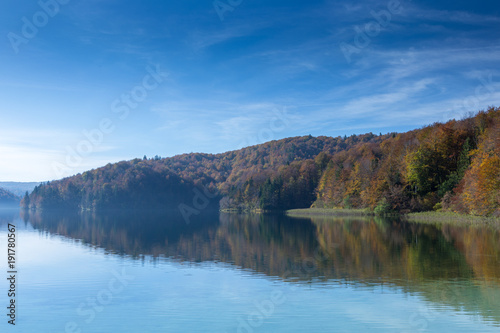 Reflection of a peninsula on a lake. Taken in Plitvice Lakes National Park. The sky is mostly clear with few clouds and the trees are turning colors.