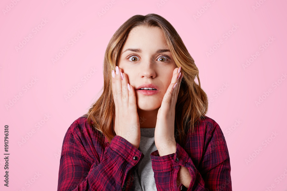 Stunned female model stares at camera, keeps hands on cheeks, being shocked with unexpected bad news, isolated over pink background. Surprised young woman says: Omg, what I see, it`s unbelievable Stunned female model stares at camera, keeps hands on cheeks, being shocked with unexpected bad news, isolated over pink background. Surprised young woman says: Omg, what I see, it`s unbelievable