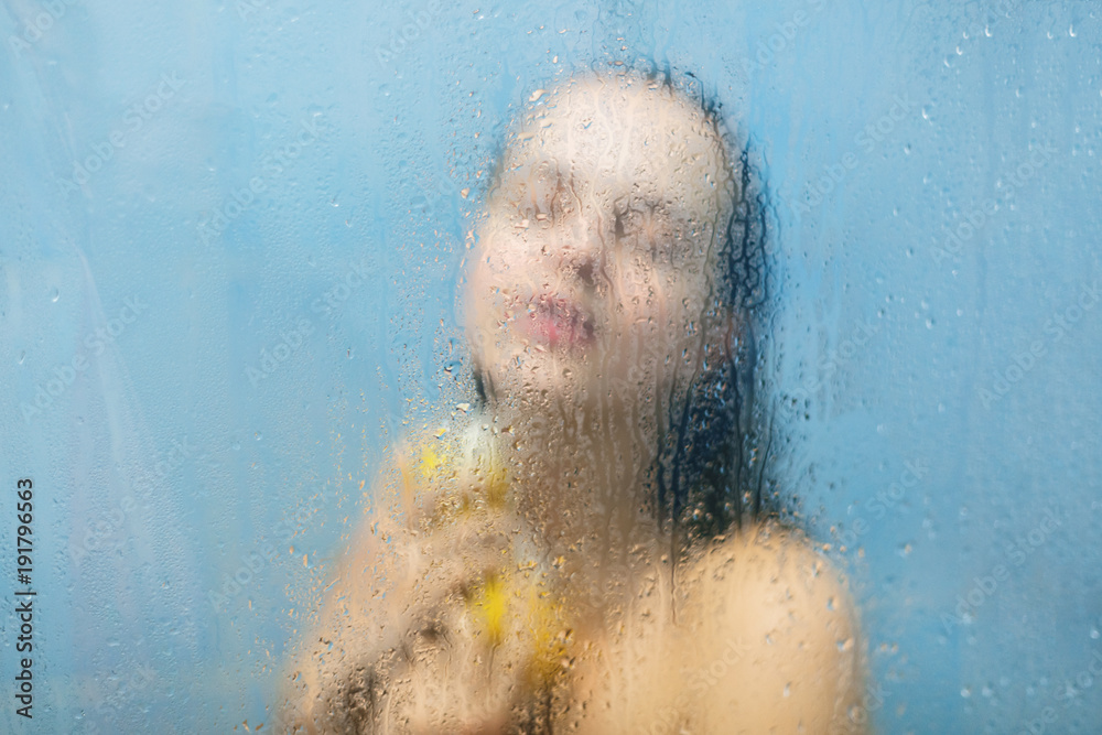 Unrecognizable female model takes shower, applies shower gel on body with bath sponge, keeps eyes shut as feels relaxed, poses in shower cabine against sweat blurred glass with drops