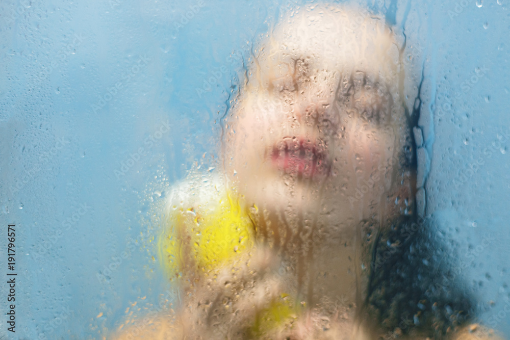 Relax in bathroom. Wet female keeps eyes closed, holds bath sponge, takes hot shower, wants to be clean and fresh, feels relaxation in shower cabine. Blurred sweat background. Wellness concept