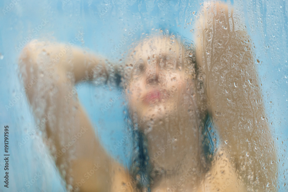 Abstract blurred background of young woman showers in bathroom, uses scrub skin and shampoo behind sweat glass with drops. Naked female under hot water in shower cabine, enjoys good relaxation