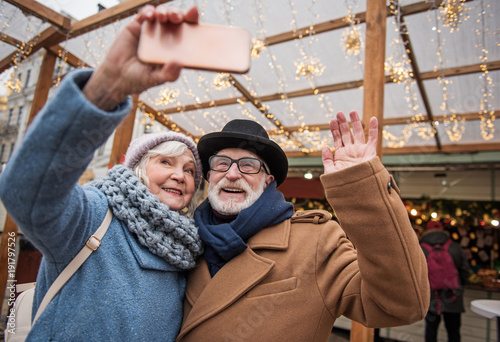 Joyful mature married couple is making selfie on street. Woman is holding smartphone and smiling. Man is waving arm to camera. Winter holiday entertainment concept