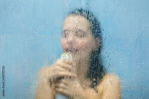 Female sings in douche, enjoys calm atmosphere and loneliness, poses naked in shower cabine, stands behind blurred background. Unrecognizable woman relaxes at bathroom. Skin care and rest concept