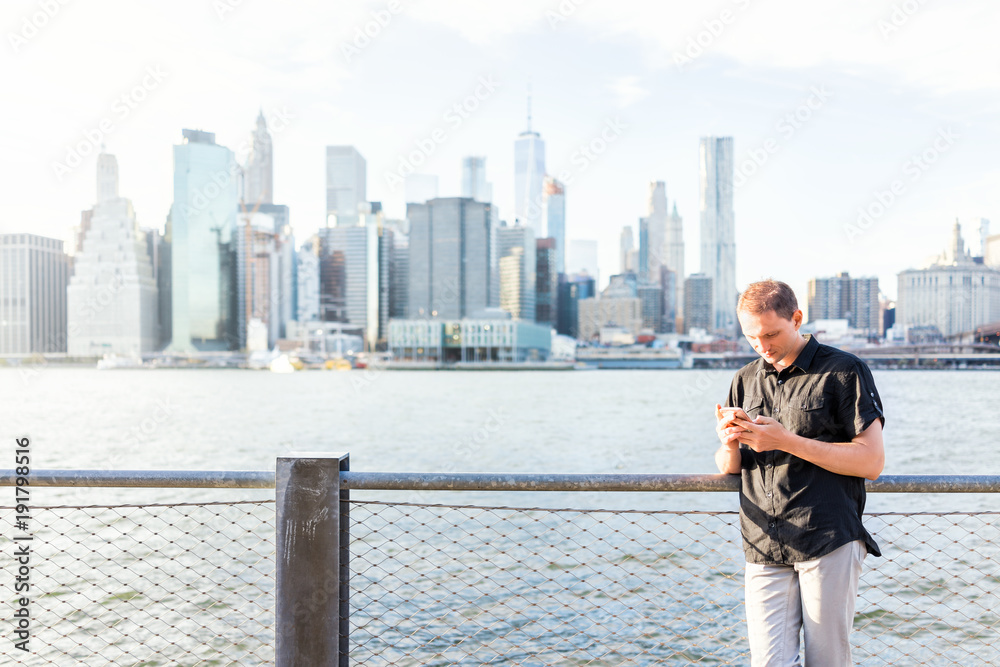 Young man outside outdoors in NYC New York City Brooklyn Bridge Park by east river, railing, looking at view of cityscape skyline, texting on social media phone