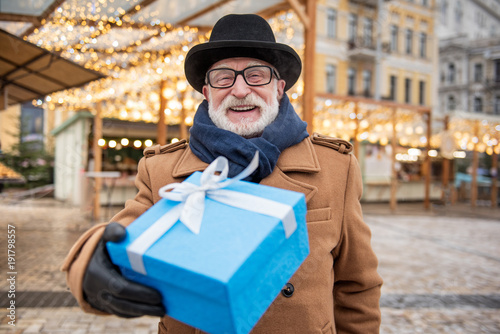 Portrait of joyful mature man stretching gift box to camera. He is standing on street and laughing. Focus on blue box