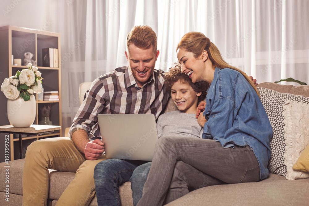 Laughing unshaven husband and beaming wife with cheerful boy watching at notebook computer. Entertainment concept