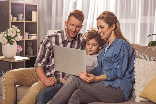 Happy bearded dad and outgoing mother with smiling son looking at laptop. They sitting on sofa. Family concept