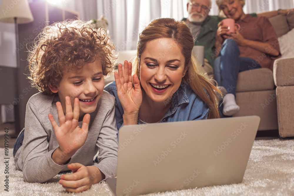 Portrait of cheerful mom and outgoing curly boy looking at laptop while lying on floor. They flourishing arms. Gadget concept
