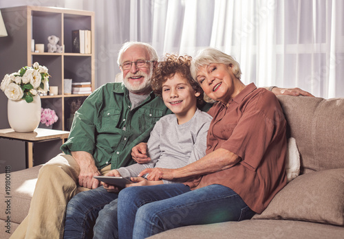 Portrait of outgoing grandfather and glad granny with friendly child watching at camera. They holding appliance on couch. Relax concept