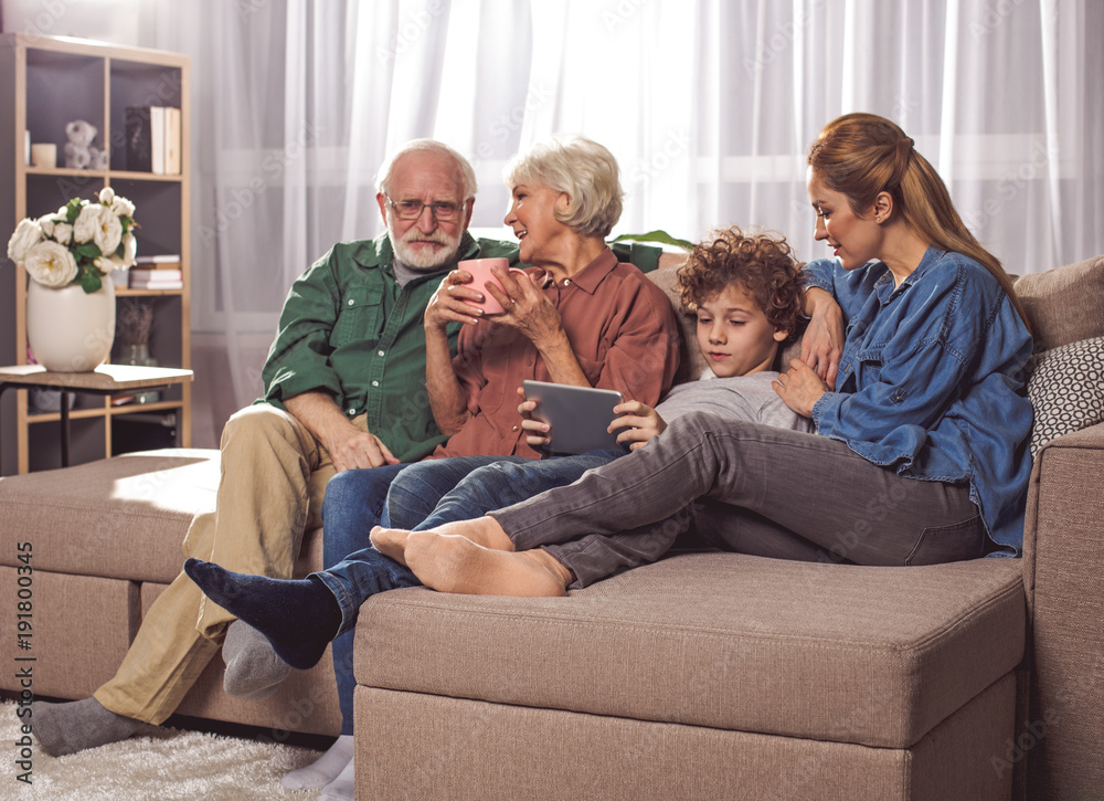 Smiling grandmother drinking cup of tea while talking with beard grandpa. Happy mom and child looking at electronic tablet. Leisure concept Smiling grandmother drinking cup of tea while talking with beard grandpa. Happy mom and child looking at electronic tablet. Leisure concept