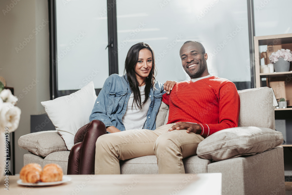 Portrait of happy man and woman relaxing on comfortable sofa at home. They are looking at camera with smile