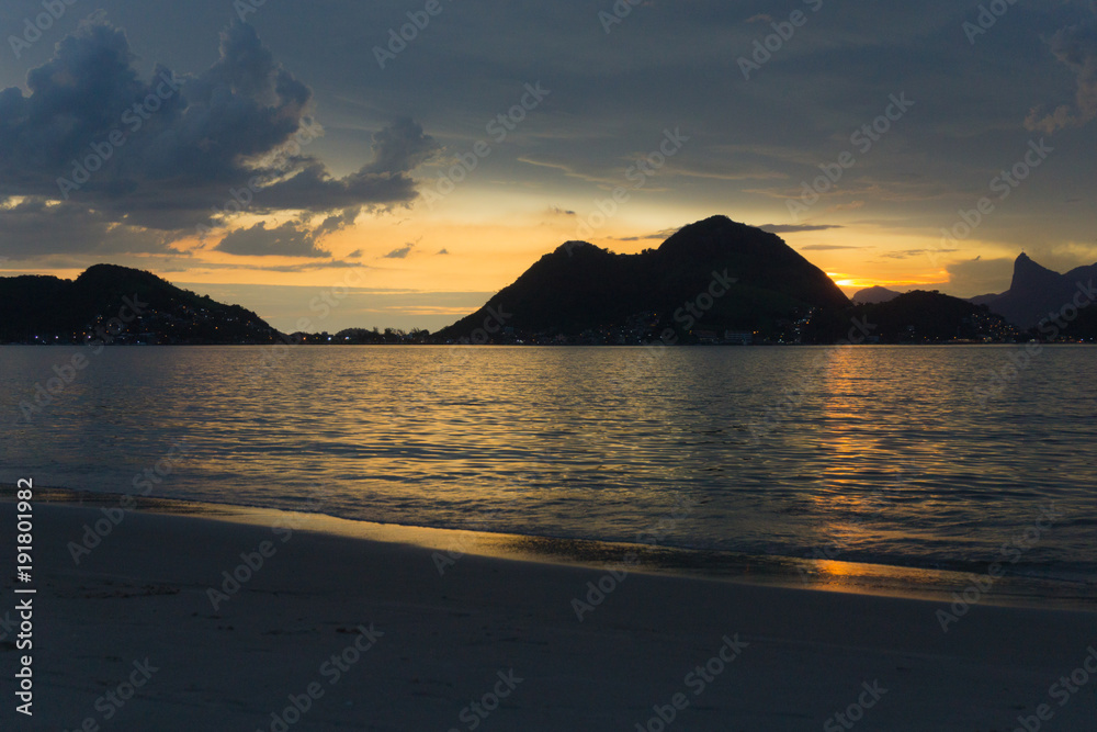 Dusk on the beach of San Francisco, Niteroi, Rio de Janeiro, Brazil. The sky is orange, the sea calm with the sun reflecting in the sea; you can see the silhouette of the mountains of the city of Rio.