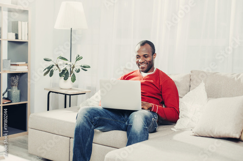 Portrait of african man holding laptop on knees while sitting on couch. He is looking at device and smiling