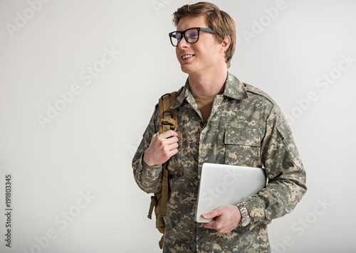 Pleased young male cadet smiling and holding laptop. He is having rucksack on shoulder and wearing eyeglasses. Isolated on background. Copy space in left side
