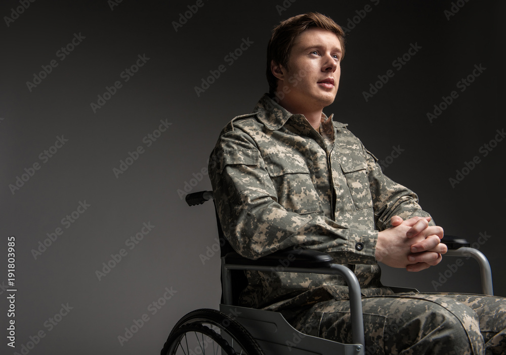 Cheerful disabled young military man looking up with hope. He is sitting in wheelchair with his hands folded in the lock. Isolated on grey background. Copy space in left side - obrazy, fototapety, plakaty 
