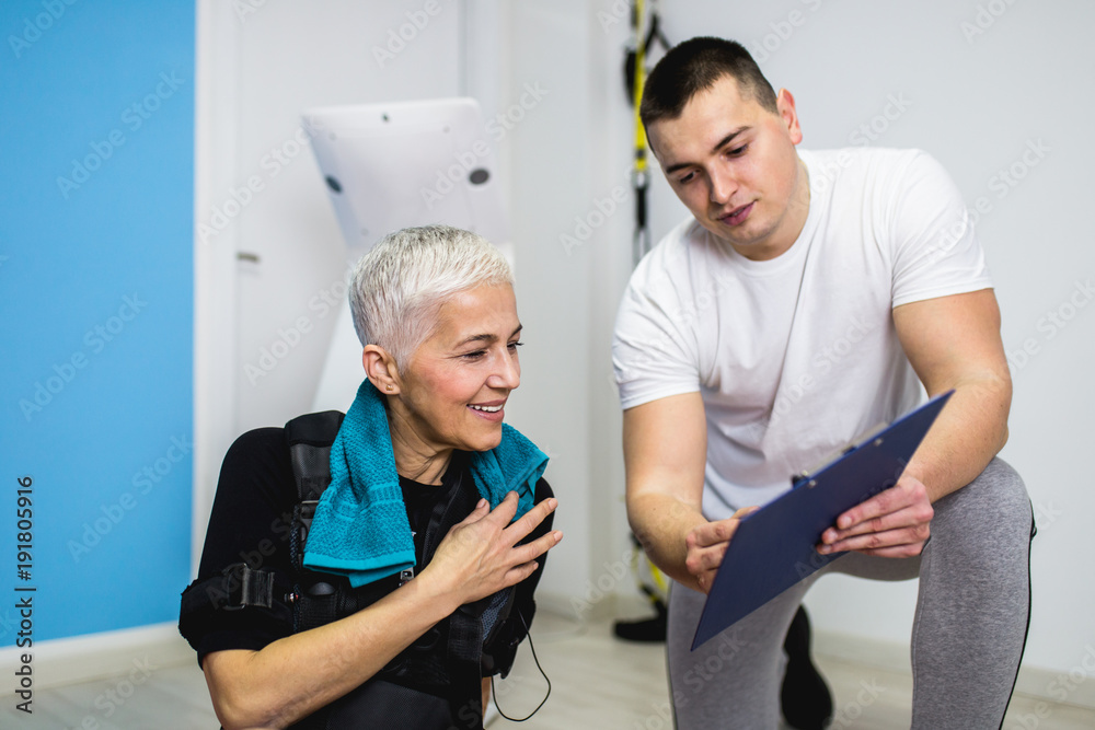 Obraz premium Beautiful senior woman doing exercises in electrical muscular stimulation suit with her personal trainer at rehabilitation center. 
