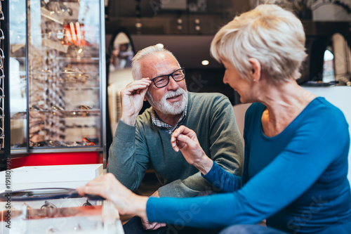 Happy senior couple choosing together eyeglasses frame in optical store. 