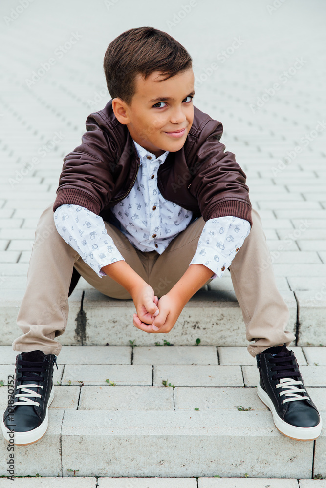 Adorable little boy sitting on stairs in a city, wearing a brown leather jacket. Adorable little boy sitting on stairs in a city, wearing a brown leather jacket.