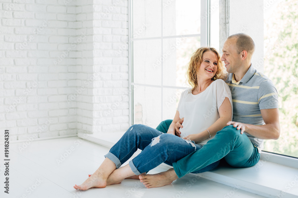 Young happy pregnant couple in love barefoot sitting on windowsill. Laughing and smiling man and woman. Happy pregnancy concept Young happy pregnant couple in love barefoot sitting on windowsill. Laughing and smiling man and woman. Happy pregnancy concept