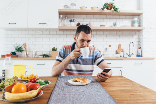 Young man sitting at table in the kitchen with a cup of coffee using mobile phone.