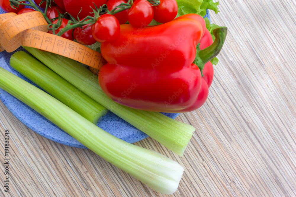 big celery,tape measure for diet, fresh green lettuce leaves next to red pepper and red cherry tomatoes lying on a blue plate, wooden table background big celery,tape measure for diet, fresh green lettuce leaves next to red pepper and red cherry tomatoes lying on a blue plate, wooden table background