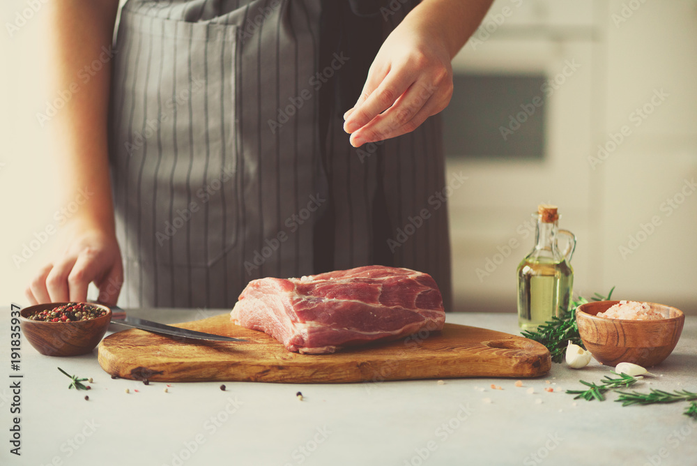 Wooman hands cutting beef meat on wooden chopping board, rosemary, oil, salt, pepper. Girk cooking pork meat on white kitchen backgound. Copy space