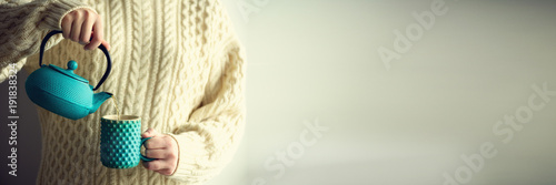 Woman in warm knitted woolen sweater holds turquoise teapot and pouring herbal tea into handmade cup. Copy space. Winter and Christmas holidays concept. Banner photo