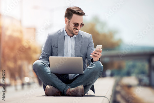 Young attractive man sitting outside on concrete while using cell phone.