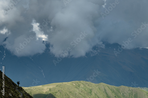 One backpaker is on a steep hillside. It looks like a small silhouette against the backdrop of large-scale mountains in the distance. Against the background of several ridges of mountains and thunderc photo