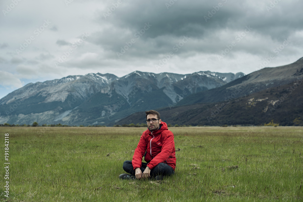 Retrato de hombre joven sentado en la verde hierba delante de un paisaje montañoso