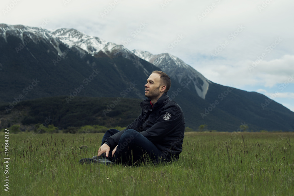 Retrato de hombre joven sentado en la verde hierba delante de un paisaje montañoso Retrato de hombre joven sentado en la verde hierba delante de un paisaje montañoso