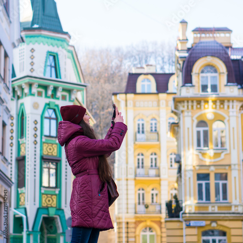 Pretty Brunette Girl Wearing Purple Winter Coat, Hat and Scarf, Walking by European Street at Winter, make Photos on Her Smartphone and Making Selfie © Oleksandr