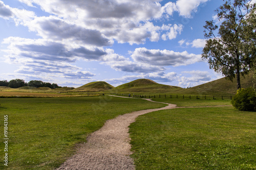 Dirt path snakes its way through the field to the royal burial mounds in Gamla Uppsala, Sweden under a blue sky with fluffy white clouds photo