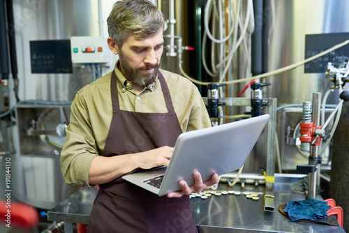 Concentrated handsome masculine middle-aged beer engineer in apron checking necessary information on laptop while working with machine at brewery factory shop