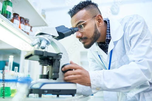 Side view portrait of young Middle-Eastern scientist looking in microscope while working on medical research in modern laboratory