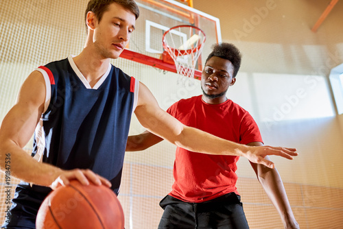 Sporty handsome young multiethnic athletes playing basketball on court: concentrated Caucasian man protecting ball from competitor while dribbling