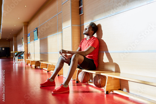 Exhausted thoughtful handsome young African-American basketball player sitting on bench with two balls under it and texting message on smartphone while stuck in training