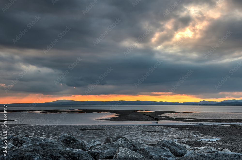 Obraz premium The sun sets on fire above the hills and mountains of the Lake District on the other side of Morecambe Bay, Lancashire, England