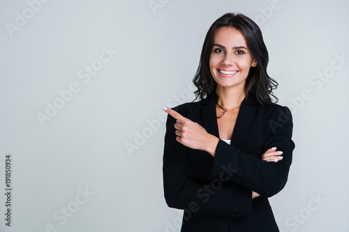 You need to look here! Beautiful young businesswoman pointing away and looking at camera with smile while standing against white background photo