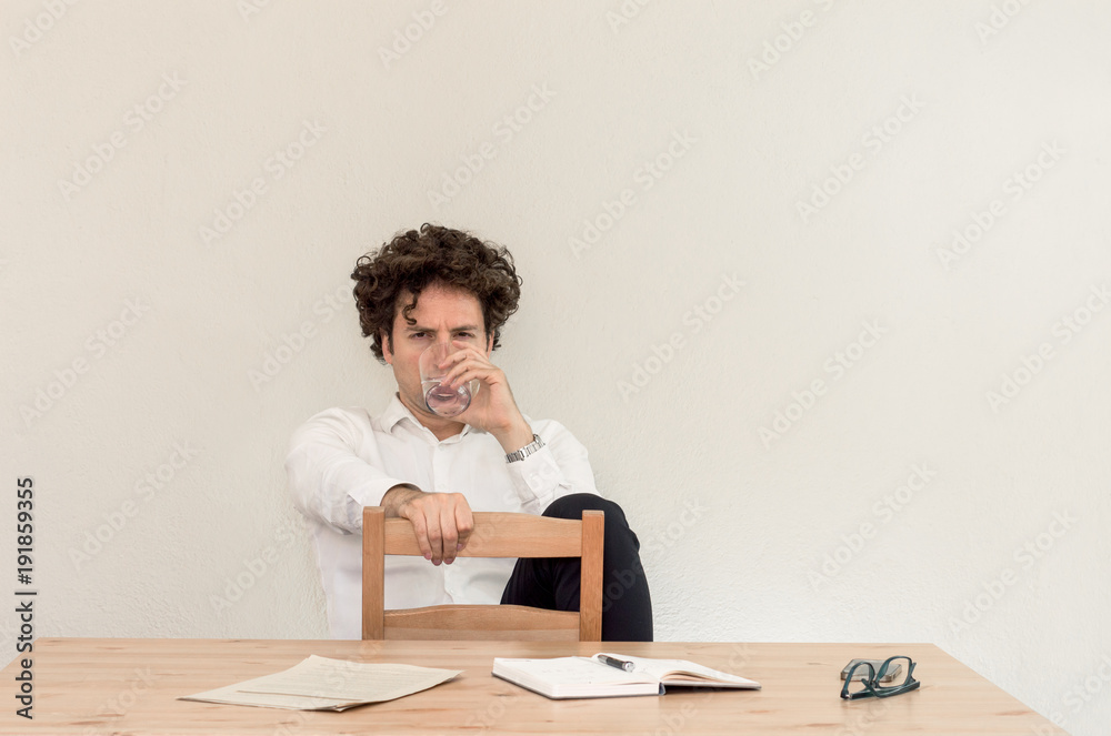 Young, Caucasian freelancer man sitting in his home office at the table, drinking water in front of empty clear wall. Young, Caucasian freelancer man sitting in his home office at the table, drinking water in front of empty clear wall.