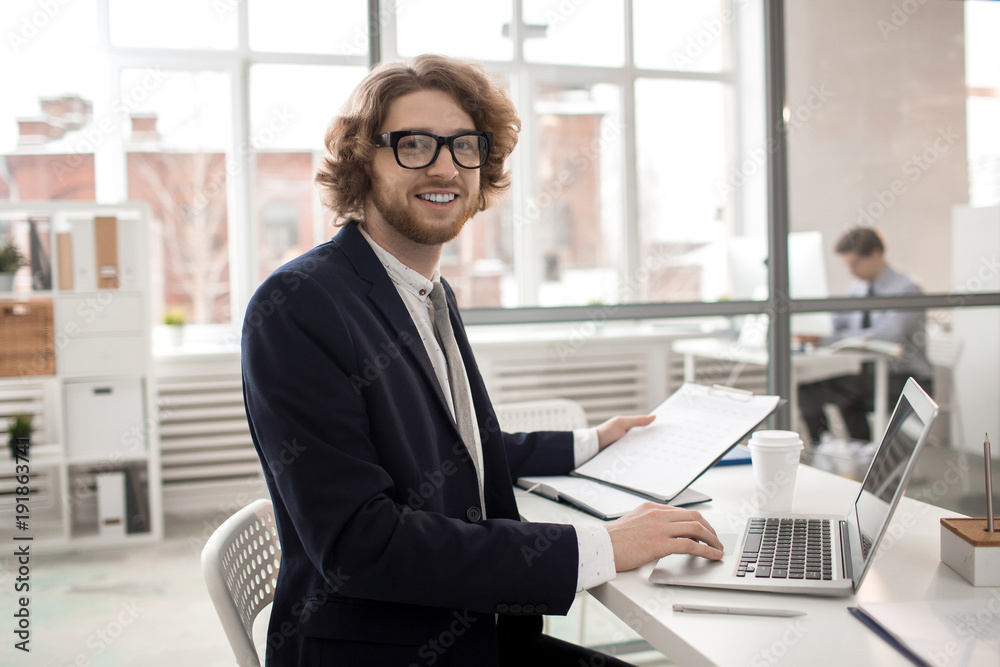 Young manager in suit and eyeglasses sitting by desk in office, analyzing online statistics and working with financial documents Young manager in suit and eyeglasses sitting by desk in office, analyzing online statistics and working with financial documents