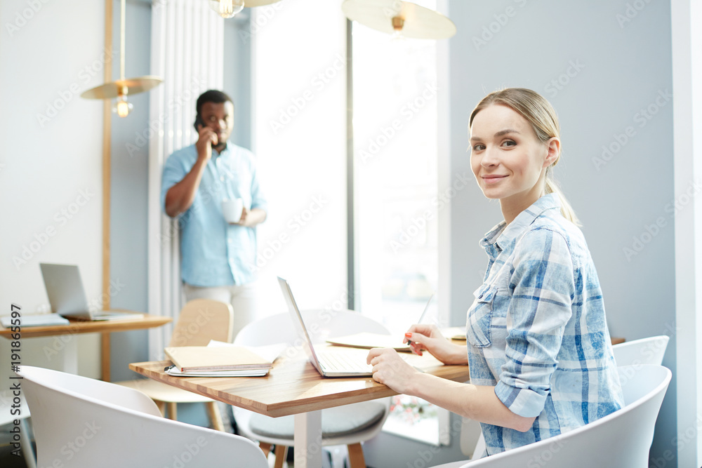 Fototapeta premium Happy young businesswoman sitting by table in office and preparing for report with man calling on background
