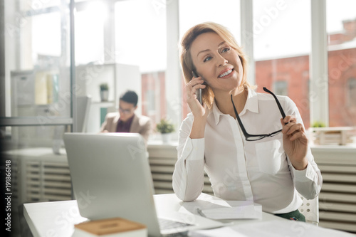 Cuadro en lienzo Smiling mid-aged woman in white shirt sitting by workplace and consulting client