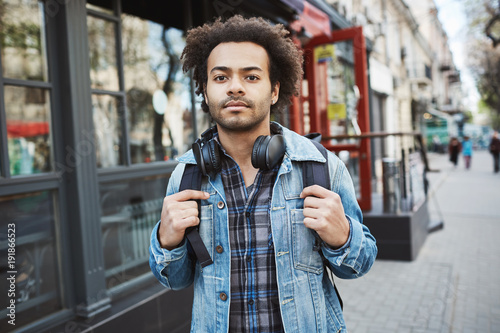 Outdoor portrait of stylish dark-skinned traveler with afro hairstyle walking on street. Man tries to find hostel where he booked a room to stay during journey to foreign country