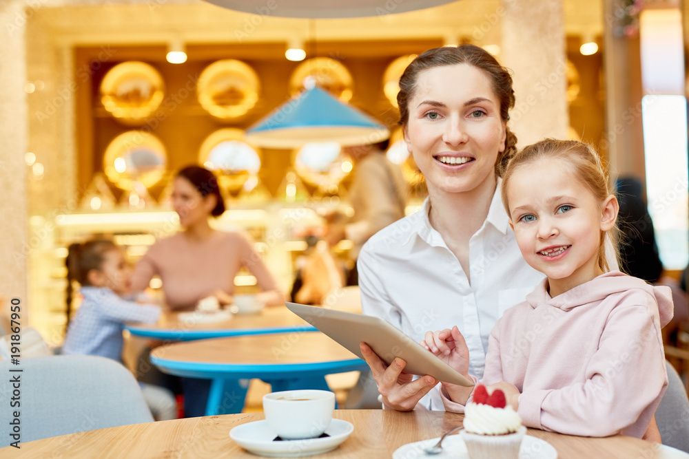 Young smiling female and her cute daughter sitting by table in cafe, having dessert with tea and watching online movie Young smiling female and her cute daughter sitting by table in cafe, having dessert with tea and watching online movie