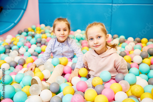Two cute little girls looking at camera while sitting among colorful balloons in theme park